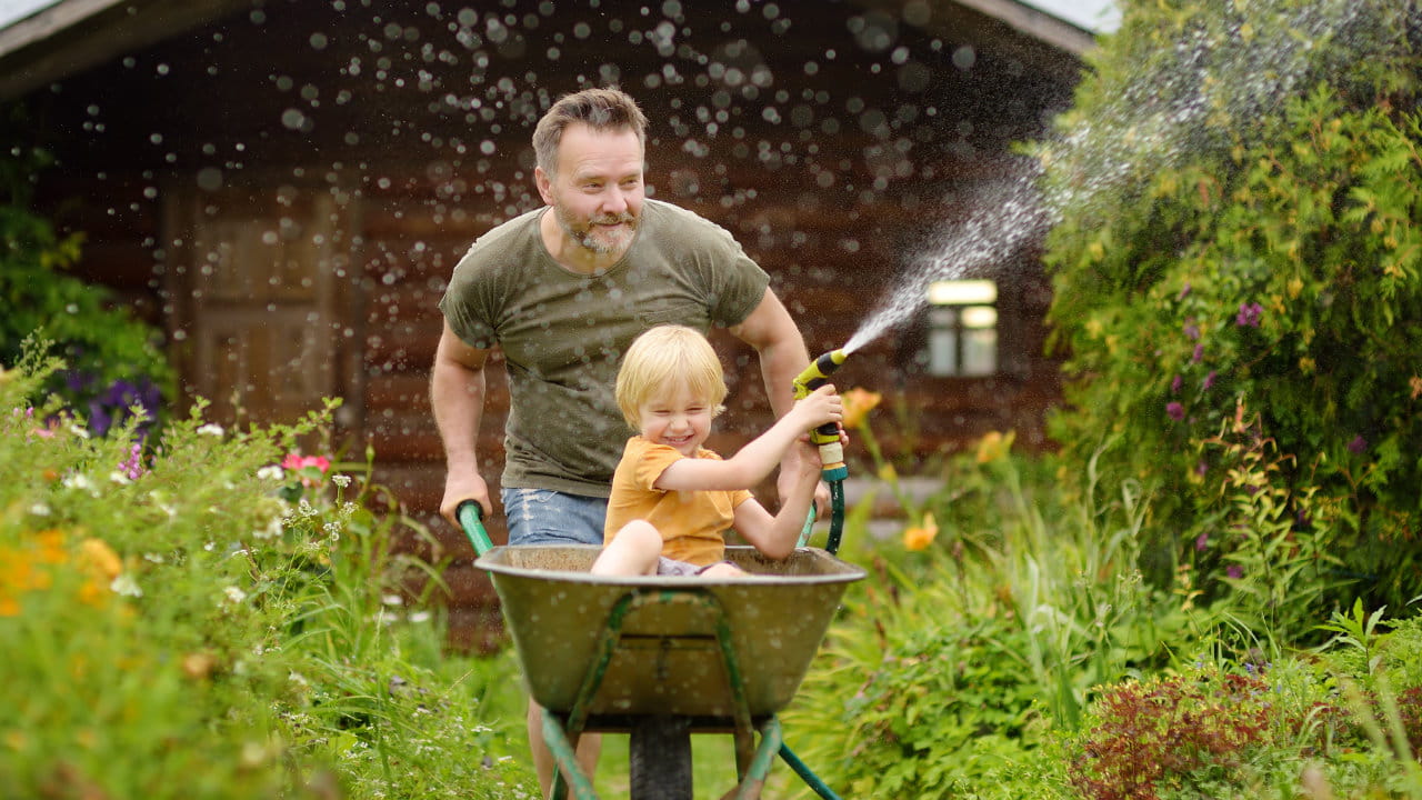 Vater schiebt ein Kind in einer Schubkarre im Garten, das mit einem Gartenschlauch Wasser spritzt