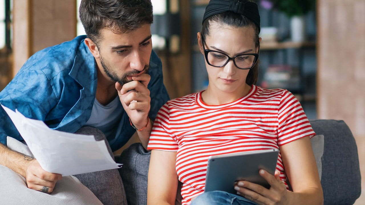 Paar sitzt auf Sofa, hält Dokumente und nutzt ein Tablet