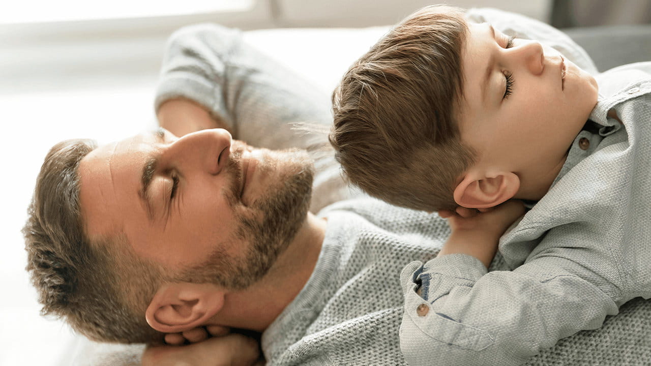 Mann liegt auf dem Rücken, Kind liegt auf ihm, beide haben die Augen geschlossen