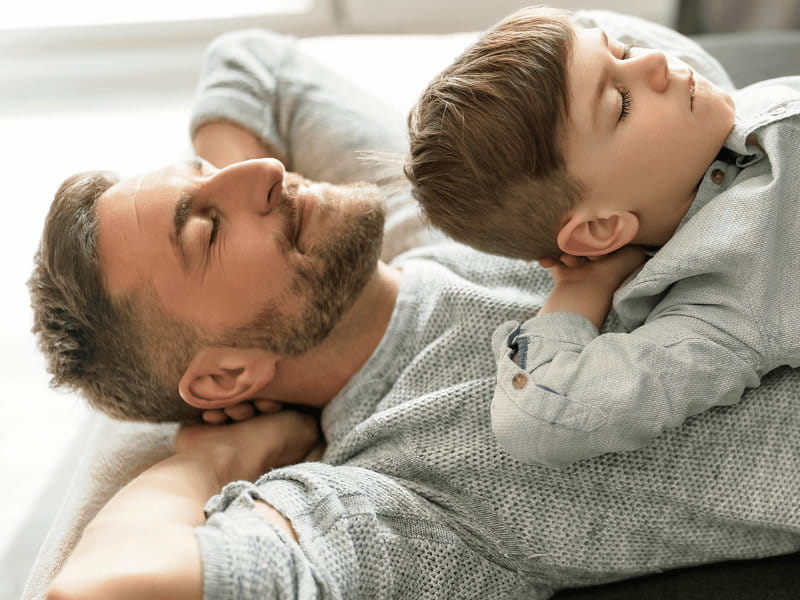 Mann liegt auf dem Rücken, Kind liegt auf ihm, beide haben die Augen geschlossen