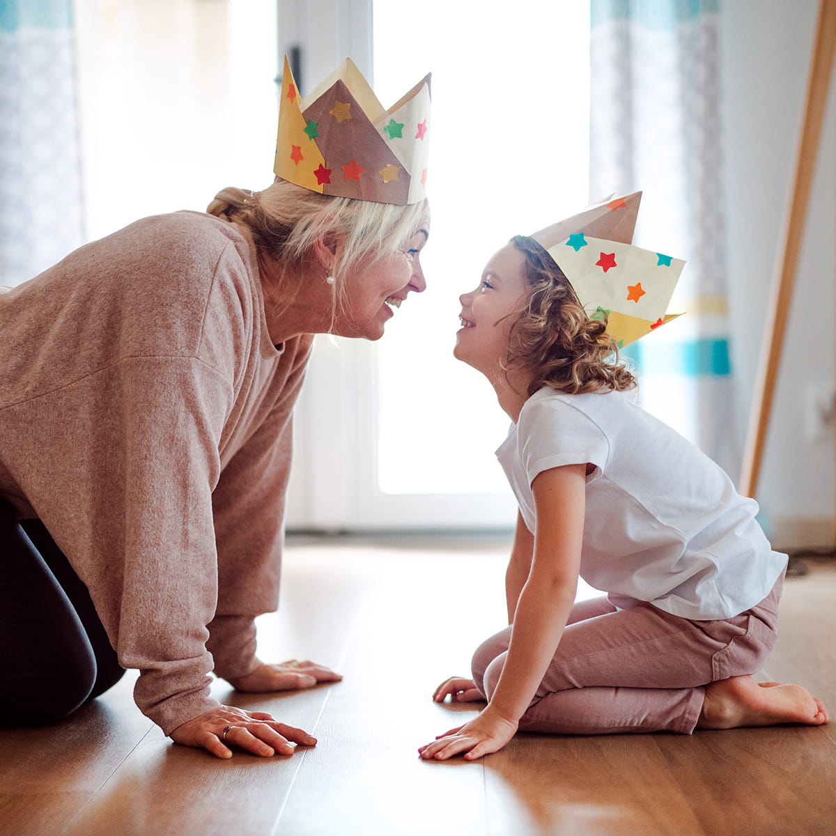 Oma und Enkelin sitzen sich gegenüber auf dem Boden und tragen Papierkronen