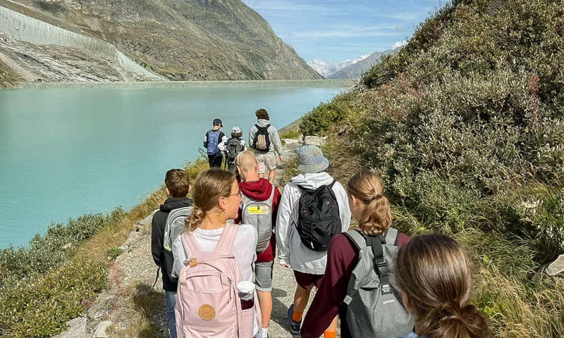 Schülerinnen und Schüler mit Rucksäcken wandern während eines Klassenlagers an einem Bergsee entlang.