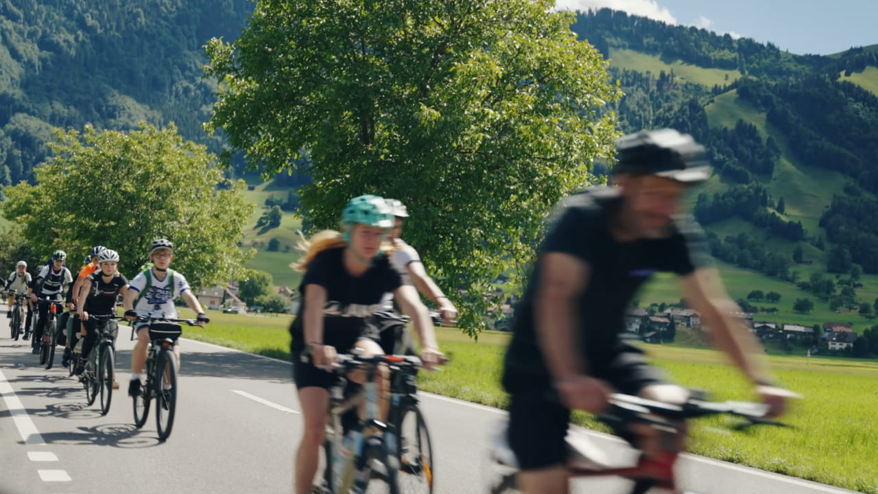 Gruppe eines Schullagers fährt mit Fahrrädern auf einer Landstrasse durch eine grüne Hügellandschaft.