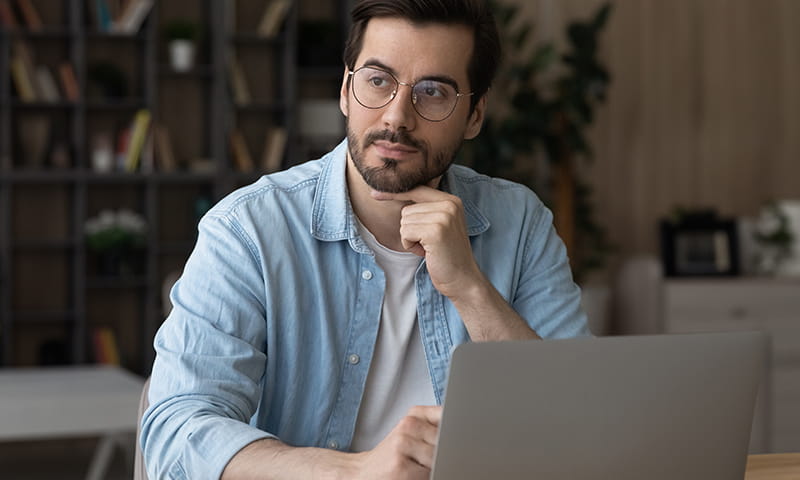 Mann sitzt am Tisch, blickt auf einen Laptop und stützt das Kinn auf die Hand