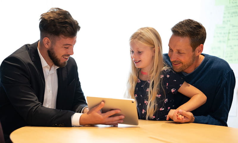 Ein Berater zeigt Vater und dessen Tochter etwas auf einem Tablet.