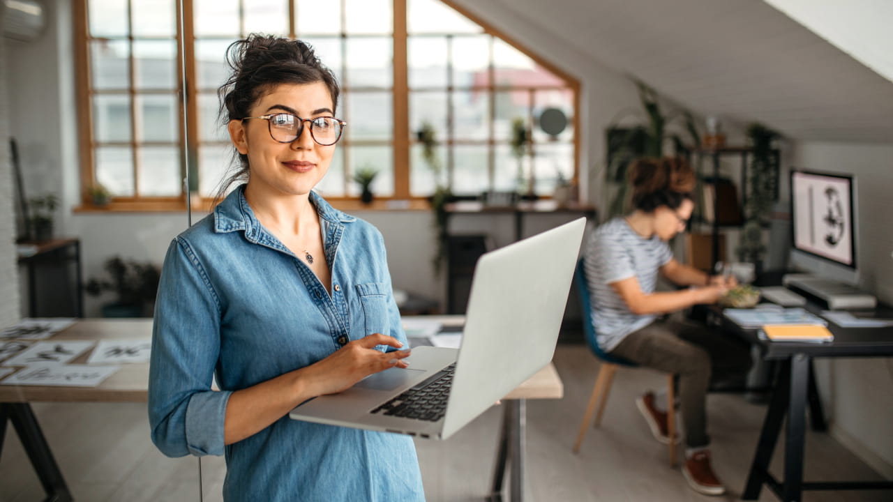 Frau arbeitet mit einem Laptop in einem Büro; im Hintergrund arbeitet eine weitere Person an einem Schreibtisch.