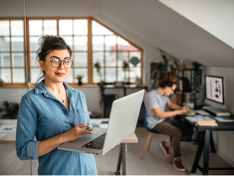 Frau arbeitet mit einem Laptop in einem Büro; im Hintergrund arbeitet eine weitere Person an einem Schreibtisch.