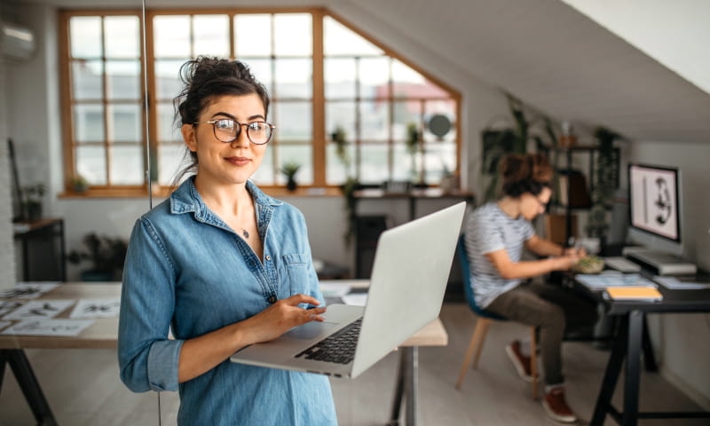 Frau arbeitet mit einem Laptop in einem Büro; im Hintergrund arbeitet eine weitere Person an einem Schreibtisch.