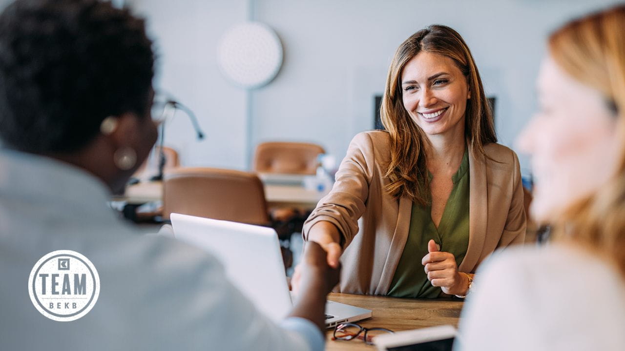 Kundenberaterin reicht einer Person die Hand bei einem Gespräch am Tisch mit Laptop